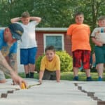 Adult with tattoos measuring material while children watch outdoors