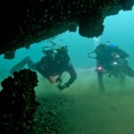 Two scuba divers exploring underwater shipwreck covered in mussels