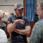 Man in baseball cap gesturing while talking to two people indoors