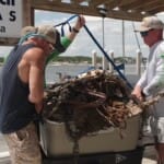 Two men lifting debris-filled container at Florida marina dock